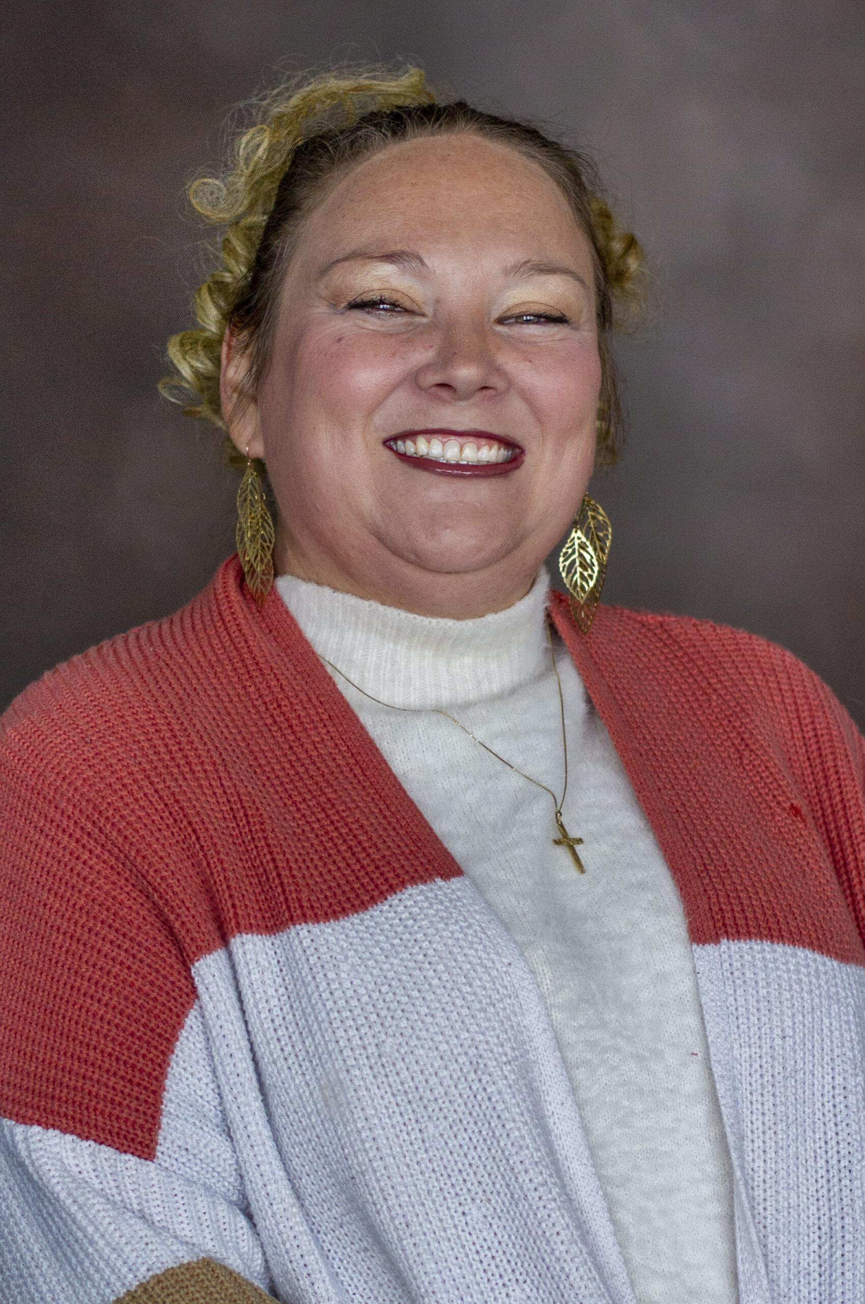 A person is smiling and wearing a red and white sweater, gold leaf earrings, and a gold cross necklace. The background is dark and neutral.